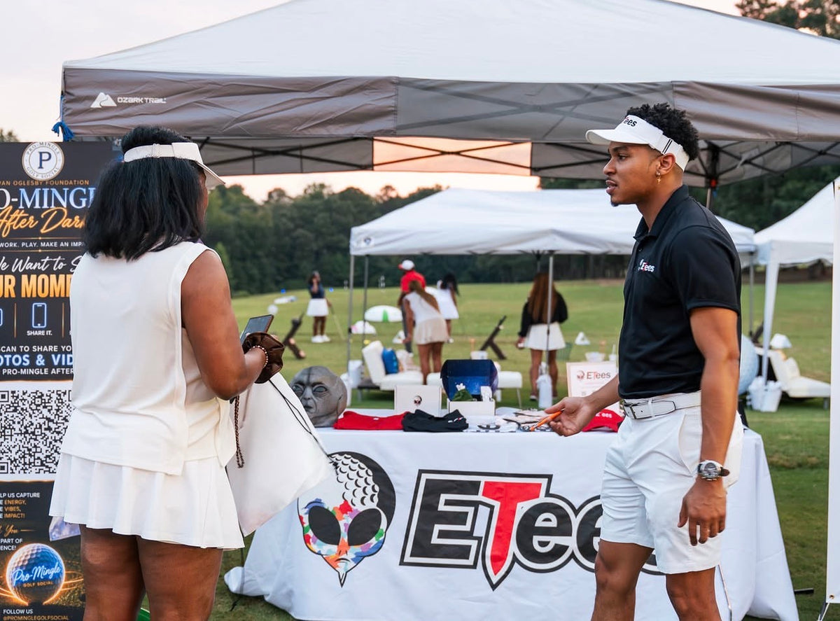A man in a black polo shirt and white shorts stands under a white canopy booth next to a woman in a white dress, with an ETees Golf table in the foreground and other event tents and people in a grassy field at dusk.